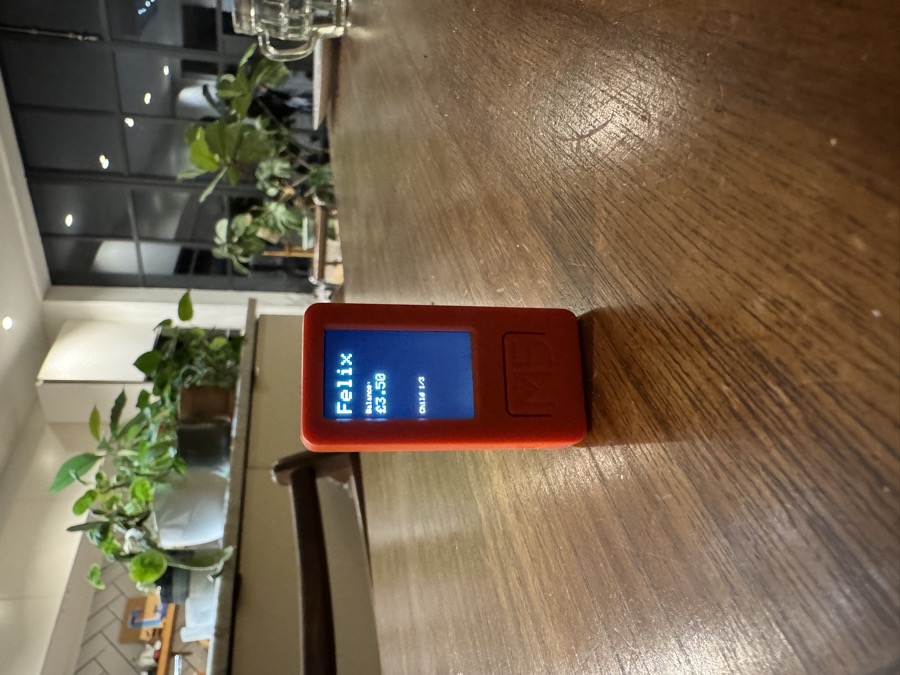 M5Stick device sitting on a kitchen table, displaying a child's bank balance of £3.50